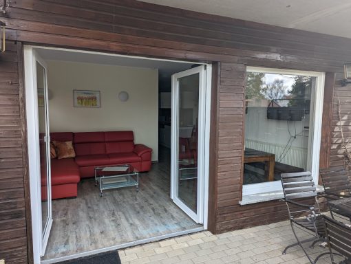 Blick aus dem Wohnzimmer des Gartenapartments Wohnbereich mit rotem Sofa, Esstisch und großen Fenstern, die auf die Terrasse blicken.